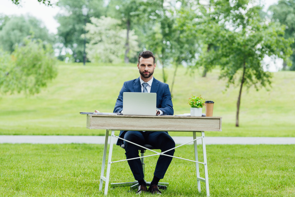 handsome young businessman in formal wear using laptop, sitting at table in park and looking at camera - Photo, Image