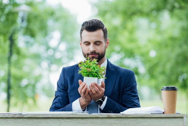 handsome young businessman holding flowerpot with plant while sitting behind table in park  - 写真・画像