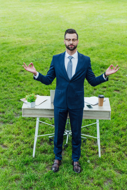 visão de comprimento total do jovem empresário em desgaste formal meditando em pé perto da mesa no parque
 - Foto, Imagem