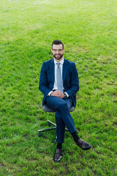 υψηλή γωνία θέα του ευτυχισμένο νεαρό επιχειρηματία κάθεται σε καρέκλα γραφείου στο πάρκο και να είναι στην κάμερα - Φωτογραφία, εικόνα