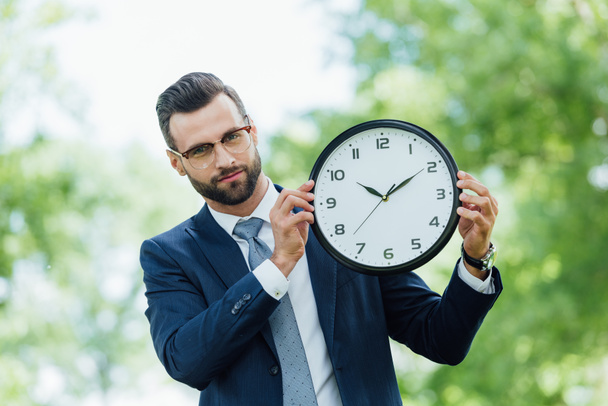 young businessman holding clock and looking at camera while standing in park - Photo, Image