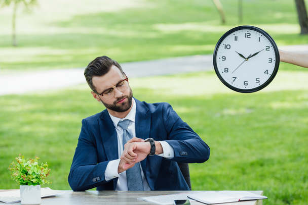 businessman sitting at table with plant in park and looking at watch  - Photo, Image