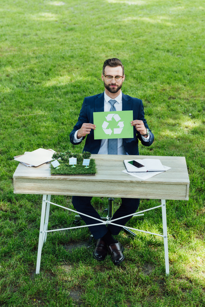 Widok z dużego kąta młodego biznesmena siedzącego przy stole z układem wiatraków, trzymający kartę ze znakiem recyklingu i patrząc na kamerę - Zdjęcie, obraz