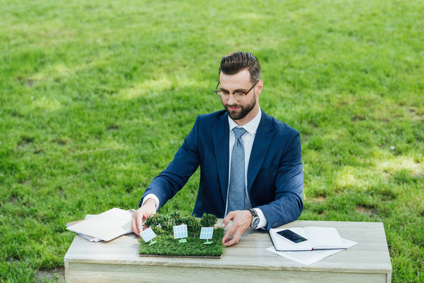 businessman putting layout with sun batteries on table with notebooks and smartphone while sitting in office chair in park  - Photo, Image