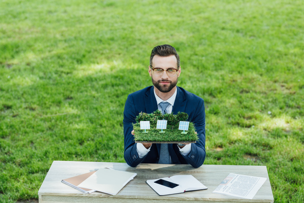 businessman holding layout with sun batteries on table with notebooks and smartphone while sitting in office chair in park  - Photo, image