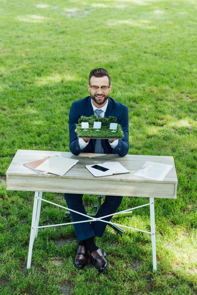 Вид бизнесмена в полный рост, держащего макет с солнечными батареями на столе с ноутбуками и смартфоном, сидя в офисном кресле в парке
  - Фото, изображение