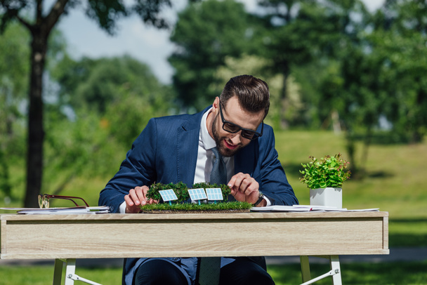 jeune homme d'affaires en lunettes de soleil assis à la table avec disposition des batteries de soleil et pot de fleurs avec plante
  - Photo, image