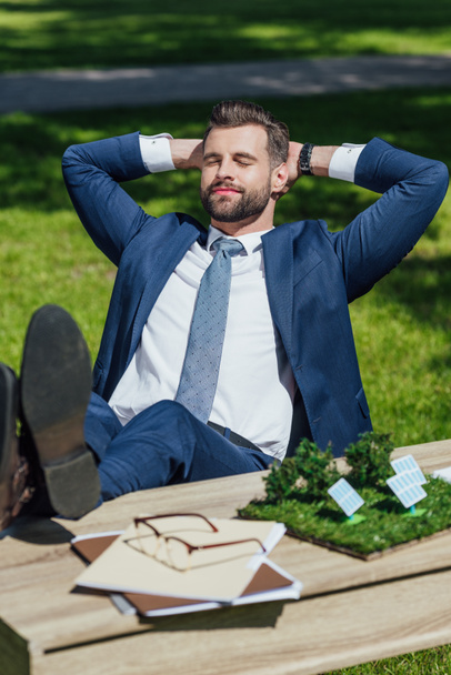 молодой бизнесмен расслабляется, сидя за столом с солнечными батареями макет, очки и ноутбуки, скрещенные лаги и положить руки за голову
 - Фото, изображение
