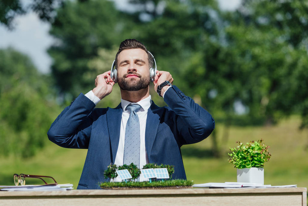 young businessman sitting at table with sun batteries layout and flowerpot, relaxing while listening to music with headphones - Photo, Image
