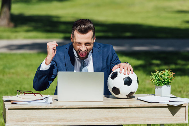 young businessman sitting at table with laptop, flowerpot and glasses, watching football, cheering on team and holding soccer ball - Photo, Image