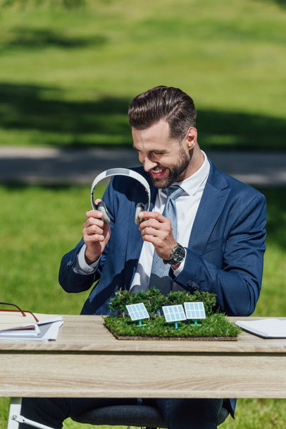 young businessman sitting at table with sun batteries layout and glasses, smiling, holding and looking at headphones  - Fotó, kép