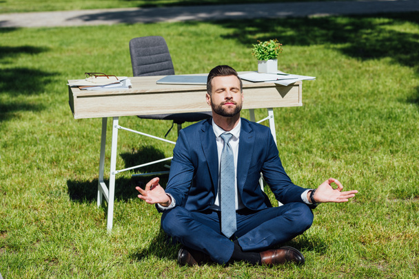 hombre de negocios sentado en la hierba y meditando cerca de la mesa en el parque
  - Foto, Imagen