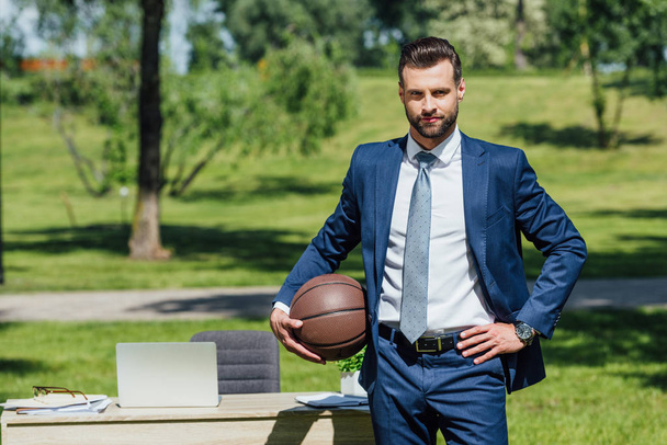 biznesmen stojący schludny stolik w parku, trzymając koszykówki i patrząc na kamery - Zdjęcie, obraz