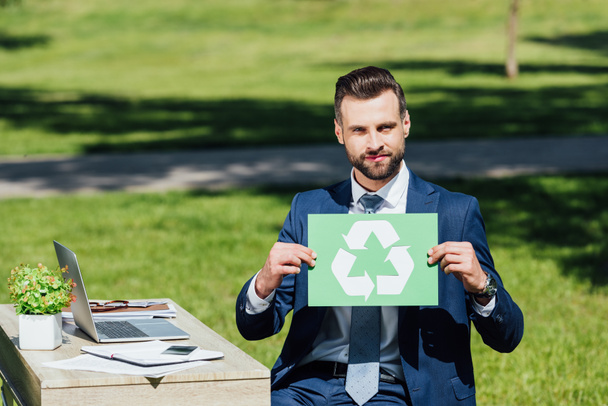 young businessman sitting in park near table with laptop, holding card with recycle sign and looking at camera - Foto, Imagen