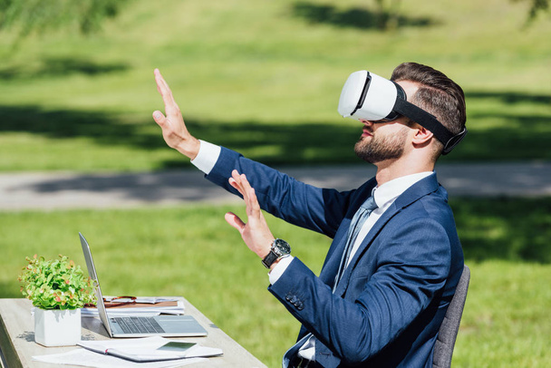 fiatal üzletember a VR headset geszturing, míg asztal, laptop, notebookok és virágcserép parkban  - Fotó, kép