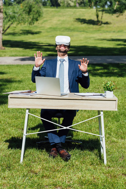 teljes hosszúságú kilátás a fiatal üzletember a VR headset gesztjáró, miközben asztal laptop, notebook-ok és virágcserép, és mosolyogva Park  - Fotó, kép