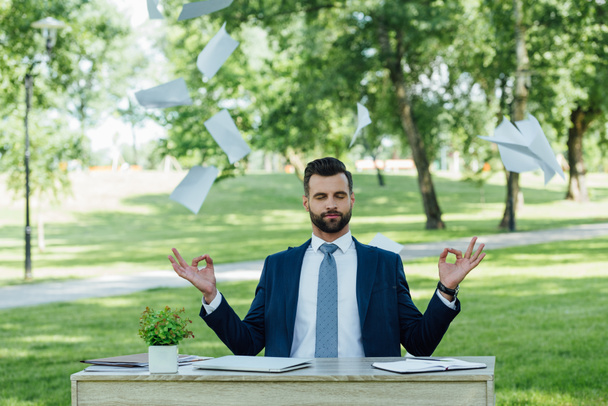 fiatal üzletember ül parkban asztal mögé, és meditál közelében repülő papírok - Fotó, kép