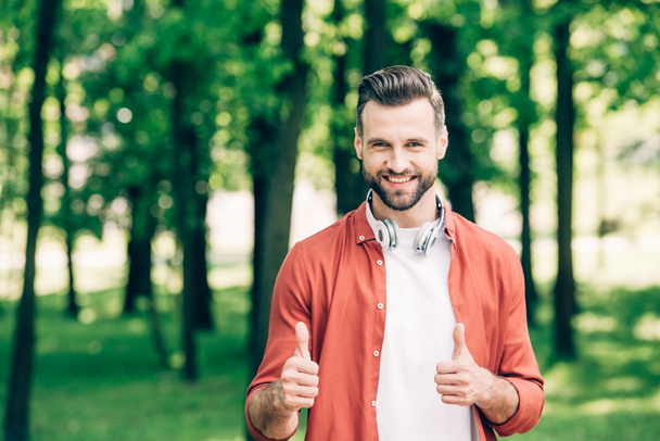 young man in red shirt standing in park with headphones on neck and showing thumbs up - Fotoğraf, Görsel