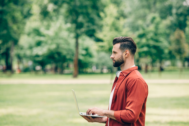 Widok z boku młodego mężczyzny stojącego w parku z laptopem w rękach i patrząc z dala - Zdjęcie, obraz