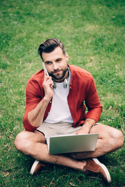 Junger Mann sitzt auf Gras im Park, telefoniert mit Smartphone und Laptop - Foto, Bild