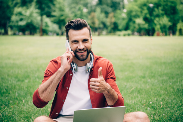 young man sitting on grass with laptop, talking on smartphone and showing thumb up - Photo, Image