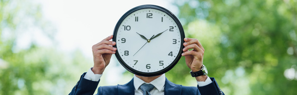panoramic shot of businessman covering face with clock while standing in park - Foto, imagen
