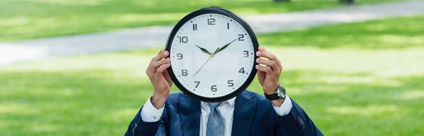 panoramic shot of businessman covering face with clock while standing in park - Fotografie, Obrázek