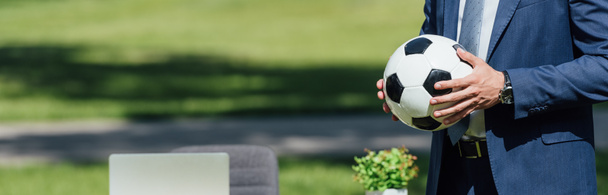 panoramic shot of businessman holding soccer ball in park near table with laptop - Fotografie, Obrázek