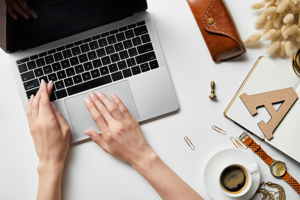 обрезанный вид женщины, работающей с ноутбуком на белой поверхности с растениями, кофе, чехлом, канцелярскими принадлежностями и блокнотом
 - Фото, изображение
