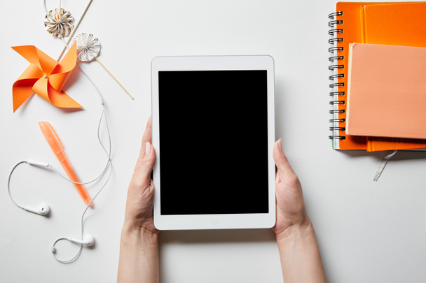 oříznutý pohled ženy držící digitální tablet v blízkosti oranžových notebooků, dekorační předměty a sluchátka na bílém povrchu - Fotografie, Obrázek