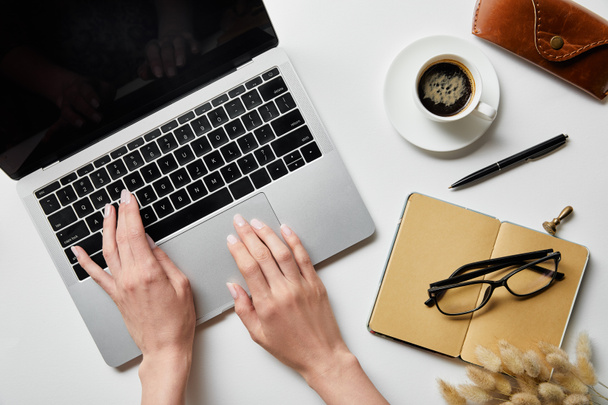 Ausgeschnittene Ansicht einer Frau, die mit Laptop in der Nähe von Brille, Kaffee, Etui und Notizblock auf weißer Oberfläche arbeitet - Foto, Bild