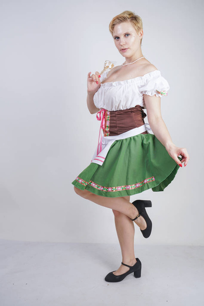 Симпатичні плюс розмір дівчини з коротким стрижка і великі груди одягнені в національну баварську сукню на білому тлі в студії. Сексуальна пишна кавказька жінка стоїть в процитувати сукню з зеленою спідницею. - Фото, зображення