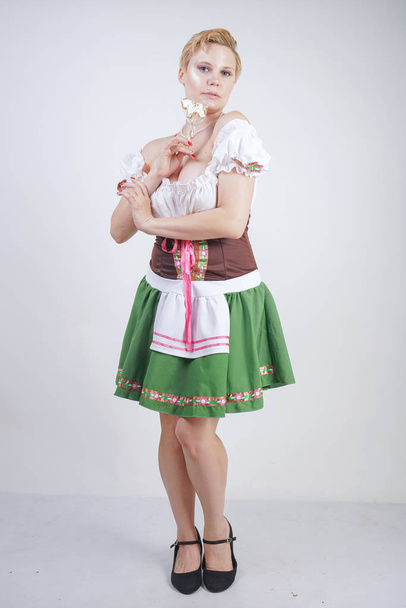 cute plus size girl with a short haircut and big Breasts dressed in a national Bavarian dress on a white background in the Studio. sexy curvy caucasian woman standing in cite dress with green skirt. - Фото, зображення