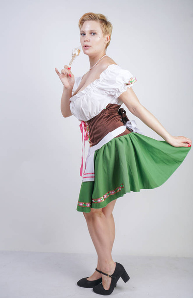 cute plus size girl with a short haircut and big Breasts dressed in a national Bavarian dress on a white background in the Studio. sexy curvy caucasian woman standing in cite dress with green skirt. - Photo, Image