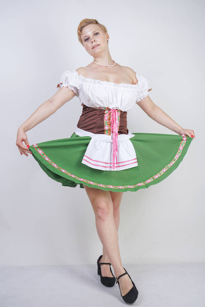 Симпатичні плюс розмір дівчини з коротким стрижка і великі груди одягнені в національну баварську сукню на білому тлі в студії. Сексуальна пишна кавказька жінка стоїть в процитувати сукню з зеленою спідницею. - Фото, зображення