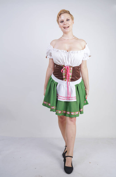 cute plus size girl with a short haircut and big Breasts dressed in a national Bavarian dress on a white background in the Studio. sexy curvy caucasian woman standing in cite dress with green skirt. - Zdjęcie, obraz