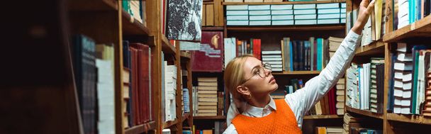 panoramic shot of pretty and blonde woman in glasses holding book in library   - Фото, зображення