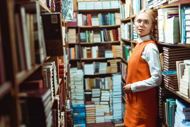 belle et blonde femme en lunettes et robe orange debout et regardant loin dans la bibliothèque
   - Photo, image