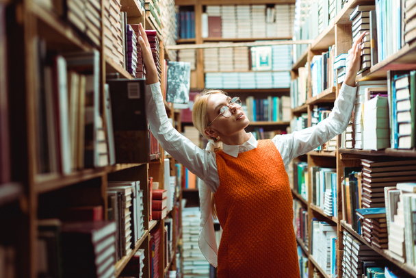 hübsche, blonde Frau mit Brille und orangefarbenem Kleid, die in der Bibliothek wegschaut   - Foto, Bild