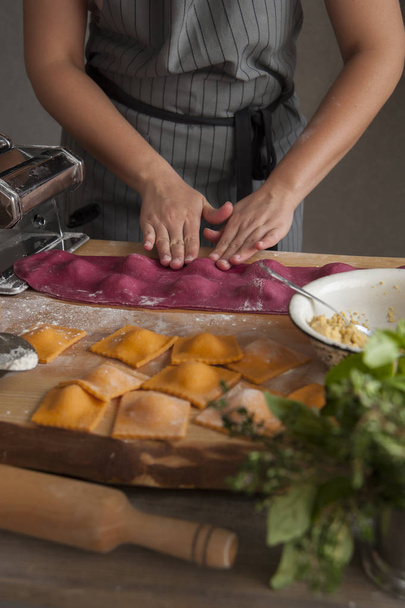 A girl in a gray apron at a wooden table makes ravioli paste from dough. In the foreground lies a rolling pin, a bouquet of herbs and cooked pasta. - 写真・画像