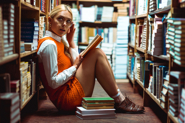 beautiful and blonde woman in glasses holding book and sitting on floor in library  - Фото, изображение