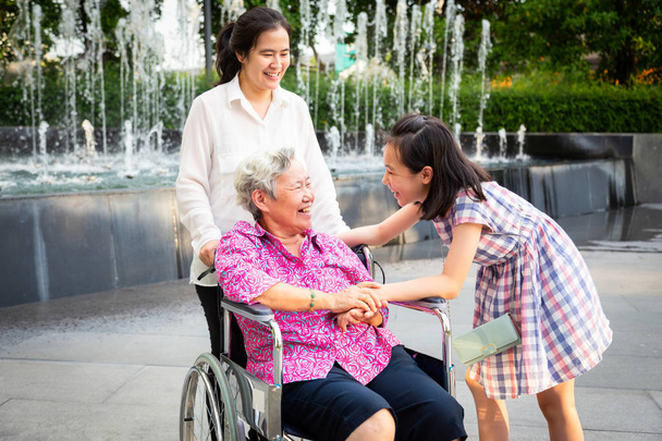 Asian senior woman having happiness and smiling with her daughte  and granddaughter on wheelchair at outdoor park,elderly woman is happy with their family,child girl having fun talk,laughing together,happy family concept - Photo, Image