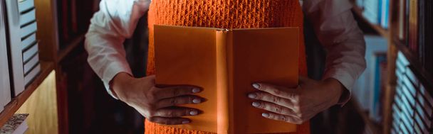 περικομμένη όψη της γυναίκας που κρατά πορτοκαλί βιβλίο με χώρο αντιγραφής στη βιβλιοθήκη  - Φωτογραφία, εικόνα