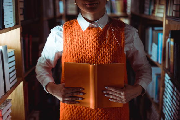vue partielle de la femme en robe tenant livre orange avec espace de copie dans la bibliothèque
  - Photo, image