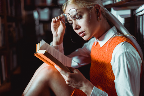 beautiful and blonde woman holding glasses and reading book in library  - Foto, Imagem
