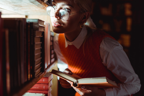 bella donna in bicchieri tenendo libro e guardando lontano in biblioteca
  - Foto, immagini