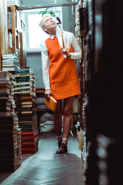 mooie en blonde vrouw in glazen en oranje jurk houden boek en opzoeken in de bibliotheek  - Foto, afbeelding