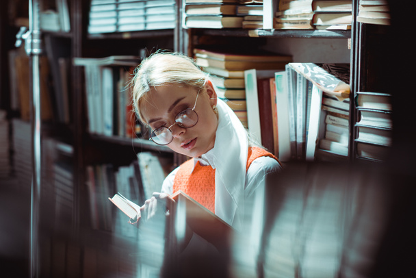 schöne und blonde Frau mit Brille liest Buch in Bibliothek  - Foto, Bild