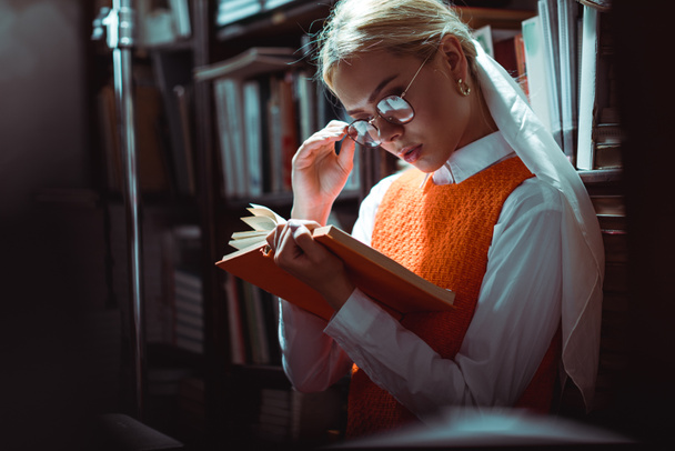 aantrekkelijke en blonde vrouw in glazen lezen boek in de bibliotheek  - Foto, afbeelding
