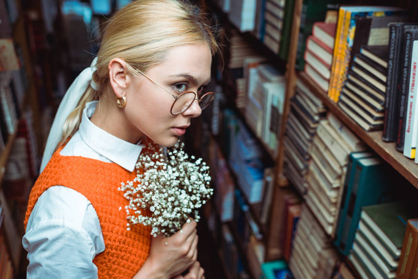 beautiful and blonde woman holding white flowers and looking away in library  - Φωτογραφία, εικόνα